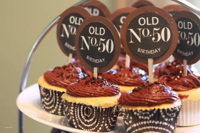 several cupcakes with chocolate frosting, decorated with round toppers, reading old No 50 birthday, in dark brown and white wrappers