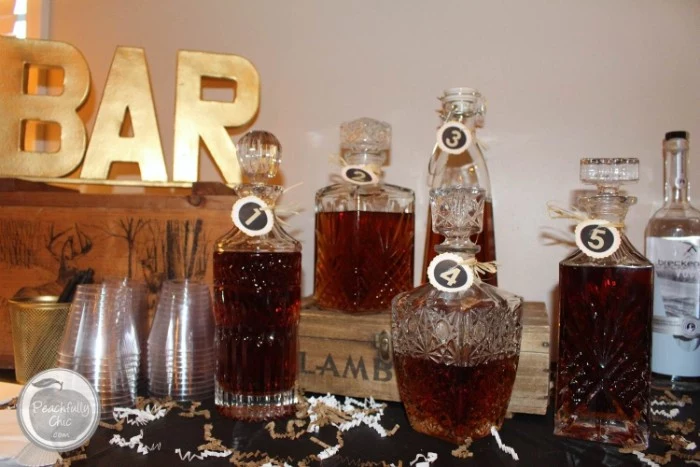 five numbered ornamental glass bottles, each containing a different kind of whiskey, placed on a table, near plastic cups, small decorative wooden crates, and a gold ornament shaped like the word bar, 50th birthday party ideas, whiskey tasting experience