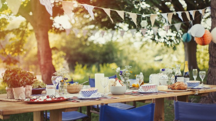 garden party 50th birthday ideas, table in a garden, set for a festive meal, several blue folding chairs, garlands and colorful paper decorations