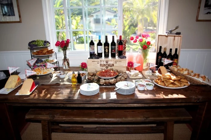 sunlit vintage wooden table, with several bottles of wine, cheese and different snacks, 50th birthday party ideas, plates and cutlery and flowers