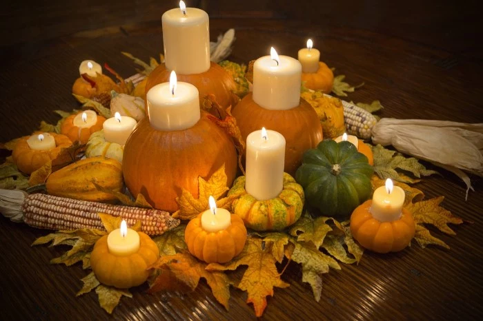 lit candles in different sizes, placed inside pumpkins, in different colors and sizes, orange and dark green, yellow with stripes, dried fall leaves, and ears of corn
