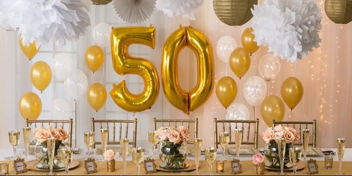 yellow and white balloons, near a table set up for a festive meal, paper decorations and lanterns, two large metallic gold balloons, shaped like the numbers 5 and 0