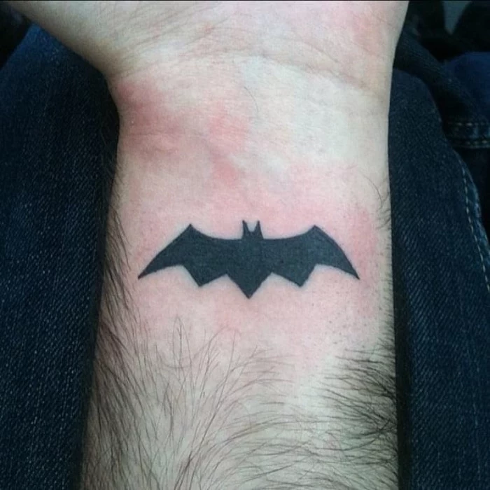 sign of batman, tattooed in solid black, on a man's wrist, lower arm tattoos, simple shape of a bat in flight