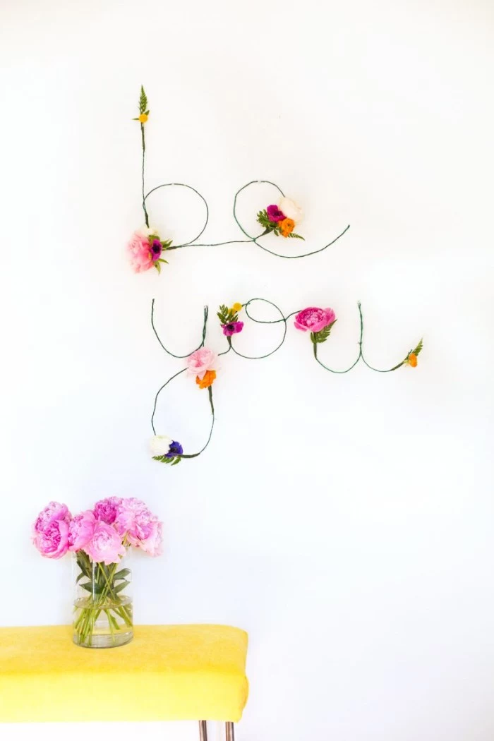 the words be you, created from green wire, and decorated with pale pink, purple and yellow faux flowers, diys for girls, mounted on a white wall, near a yellow table, with a vase, containing pink flowers