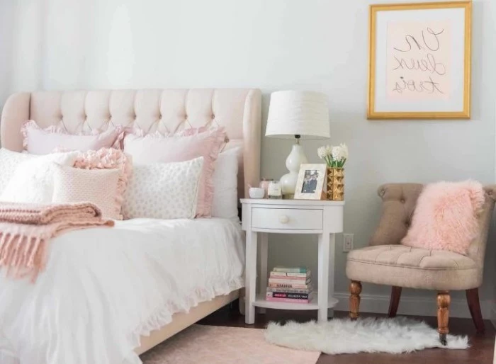 powder pink soft headboard, on a bed with white bedding, and pale pink cushions, room with light grey walls, beige vintage chair, and a white bedside table