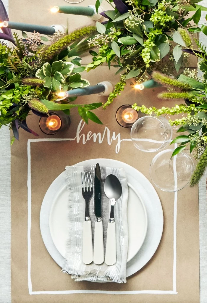 thanksgiving table setting, beige paper place mat, featuring the name henry, written in white, plates and cutlery, glasses and lit candles, three bouquets with green plants 