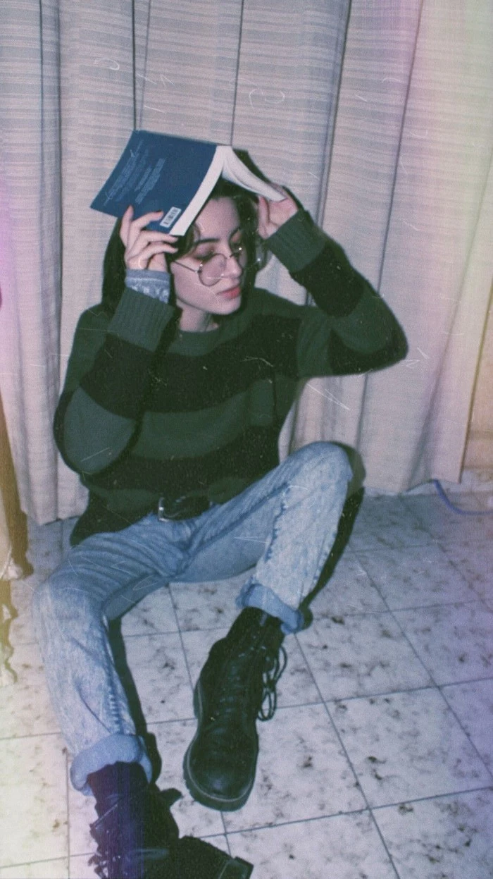pale young woman, holding an open book over her head, dressed in pale blue, acid wash jeans, and a jumper with large black, and dark green stripes, 80s grunge, black leather lace up combat boots