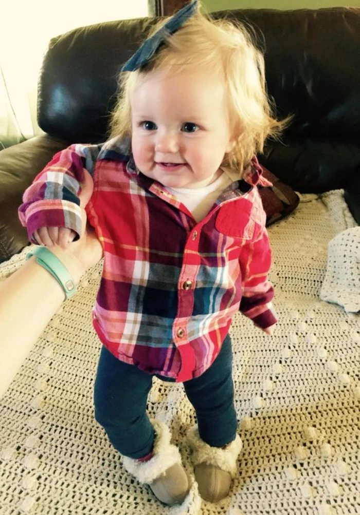 laughing one-year-old with short blonde hair, wearing dark blue skinny jeans, and a plaid shirt in red, white and dark grey, baby girl thanksgiving outfit, dark blue bow, on top of her head