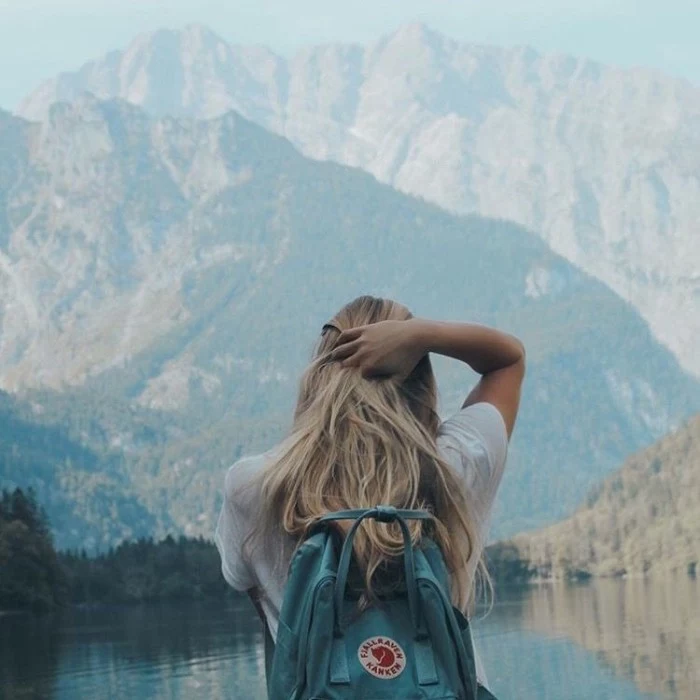 woman wearing a white t-shirt, and a teal blue backpack, running a hand through her blonde hair, while looking at a lake near a mountain, 90s grunge fashion