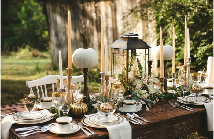 garden thanksgiving table setting, dark brown wooden table, decorated with pumpkins, a lantern filled with flowers, and several tall candles, plates and cups, cutlery and white napkins