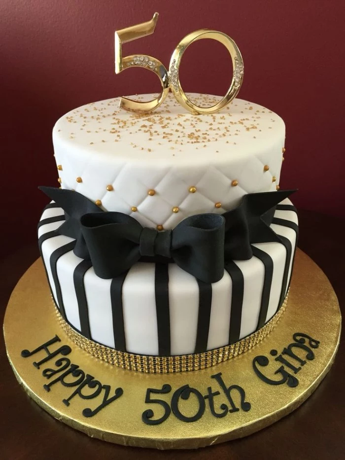 happy 50th gina, written in black on a white and gold cake, decorated with a black bow made from fondant, edible gold pearls and glitter, 50th birthday party ideas for mom, topper shaped like the number 50