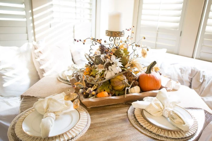 very bright room, with white window shutters, and a white sofa, near a small pale beige, round wooden table, set for two, and decorated with a centerpiece, featuring small pumpkins, gourds and leaves, berries and white flowers