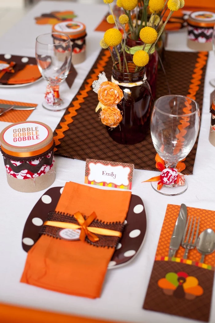 orange and brown plates, napkins and tablecloth, on a white table, decorated by a dark brown vase, containg yellow flowers, thanksgiving table setting, for the children's table