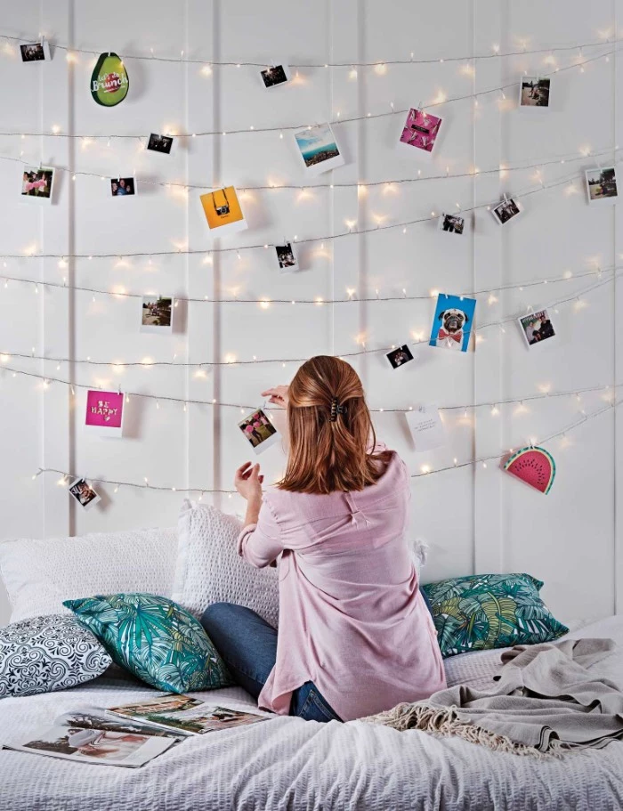cheap ways to decorate a teenage girl's bedroom, slim brunette woman, hanging a photo on a string of fairy lights, using a small clothes peg, more lit string lights overhead