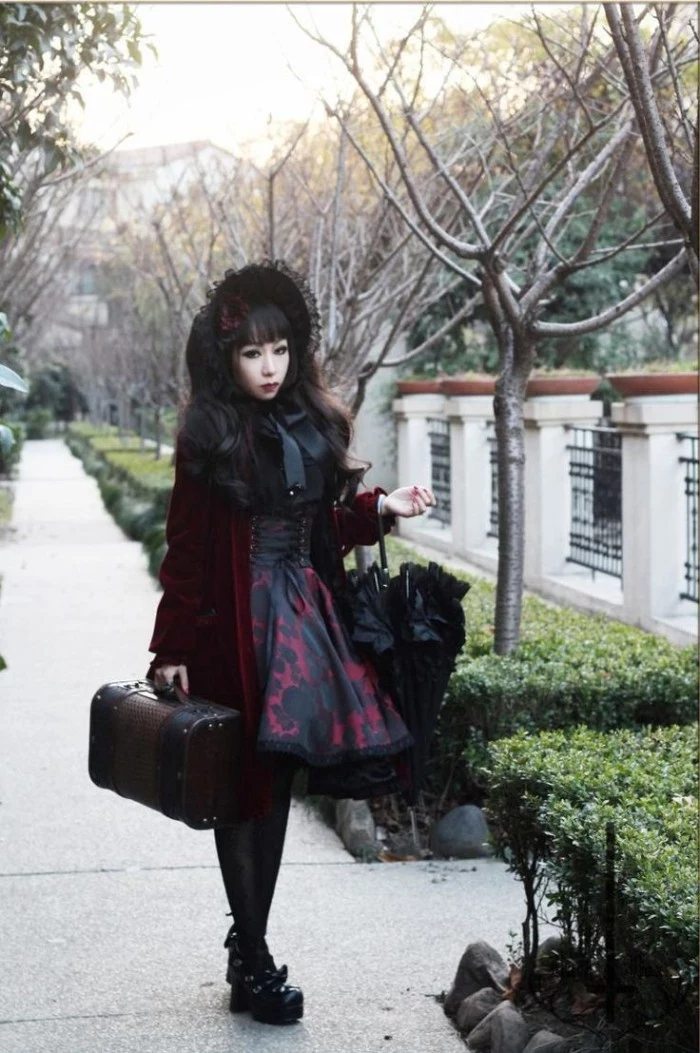 parasol in black, and a coffer bag, carried by a gothic style lolita, with a black lace bonnet, dressed in a wine red velvet coat, over a black and red dress, featuring a corset