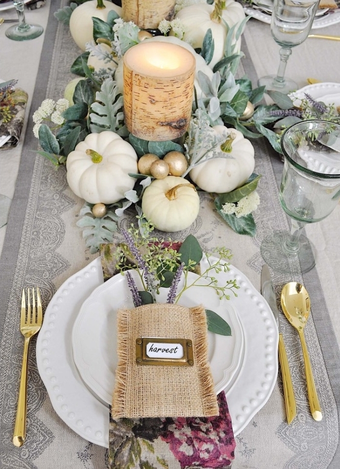 pouch made of burlap, with a small name tag, placed on top of two white stacked plates, surrounded by gold cutlery, thanksgiving table setting, candles surrounded by leaves, small white pumpkins, and gold baubles