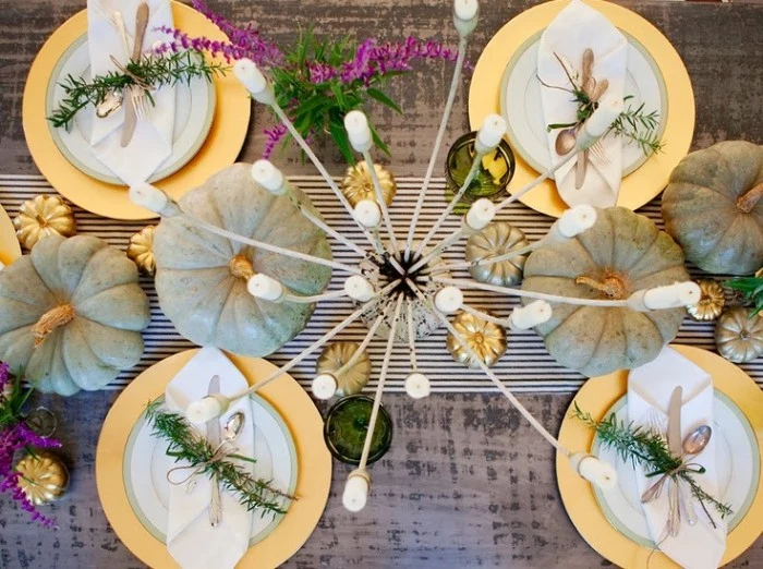 multiple white candles, in an a candle holder, placed near four pale grey pumpkins, on a table with a grey tablecloth, thanksgiving tablescape, with white and gold dishes, white napkins and silver cutlery