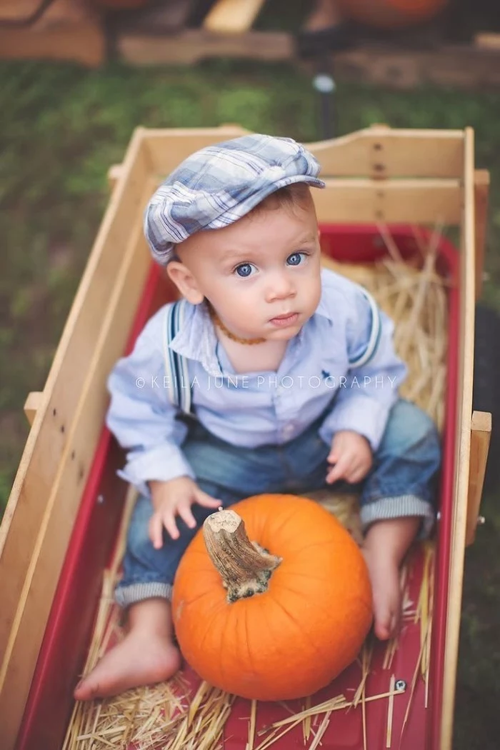 newsboy cap in white and blue, worn by a small baby boy, dressed in jeans with suspenders, and a pale blue shirt, baby thanksgiving outfits, sitting in a wooden cart, with straw and a pumpkin