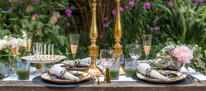50th birthday party ideas, table with colorful plates, napkins and half-filled champagne flutes, decorated with flowers and two gold candle holders, in a garden with multiple green plants