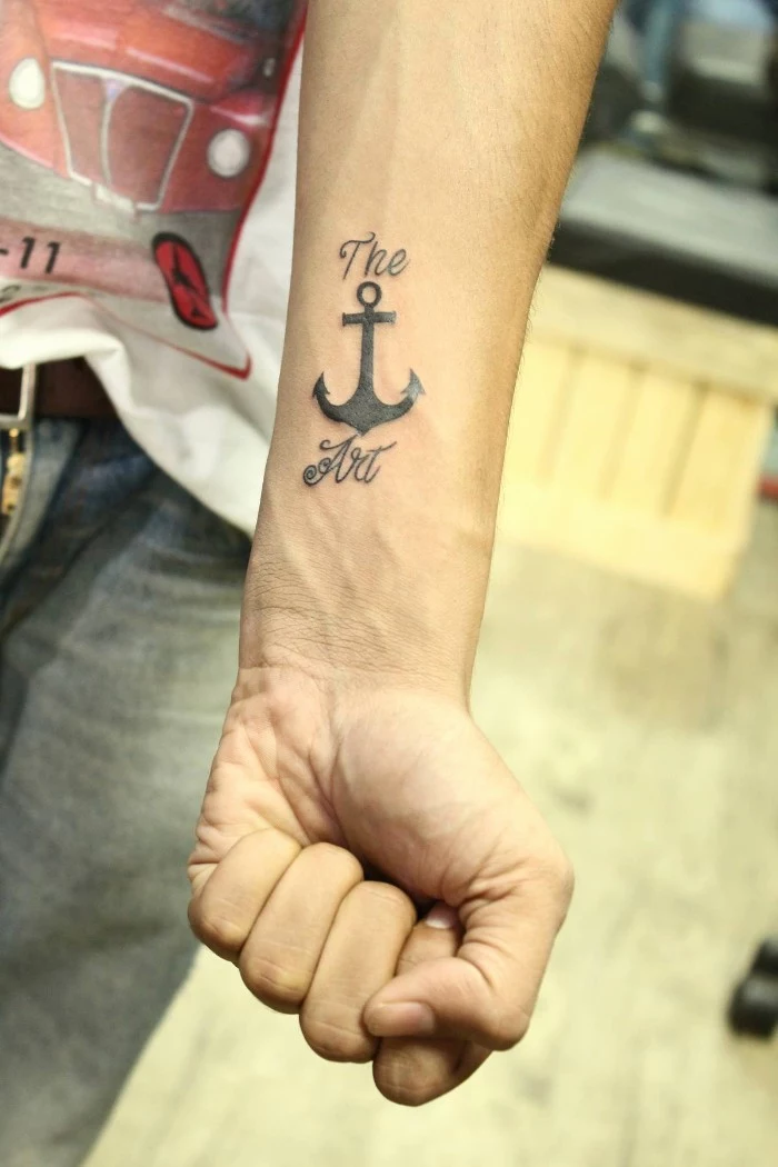 anchor tattoo in black, surrounded by the words the and art, on the lower arm of a slim man, small tattoos for men