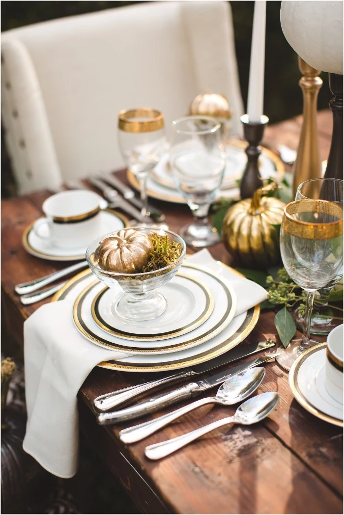 thanksgiving table decorations, spray painted gold ornamental pumpkin, placed in a glass bowl, decorated with moss, on top of three stacked plates, with a white napkin between them