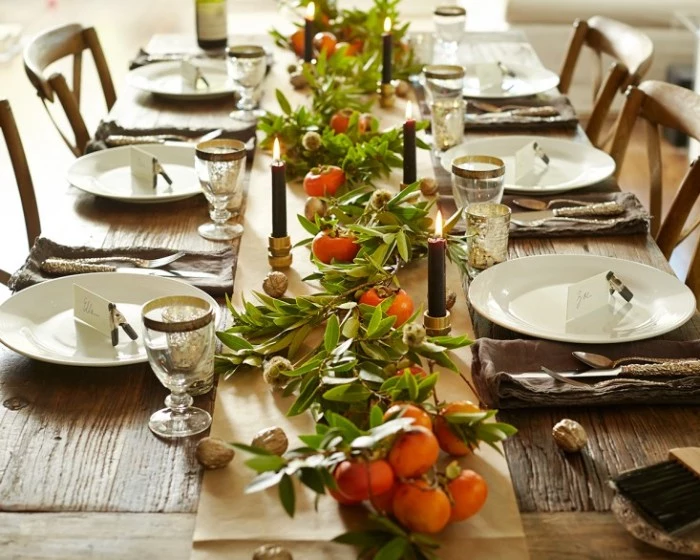 rustic style table, decorated with clementine leaves and fruit, with thanksgiving dinnerware, white round plates, and gold rimmed glasses, lit dark green candles 