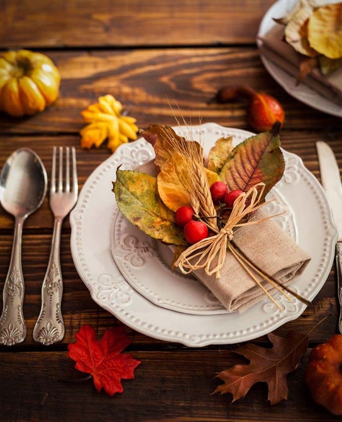 napkin made of burlap-like material, decorated with fall leaves, red berries and two sprigs of wheat, tied with paper rope, and placed on a white plate, thanksgiving table setting, silver vintage-style cutlery