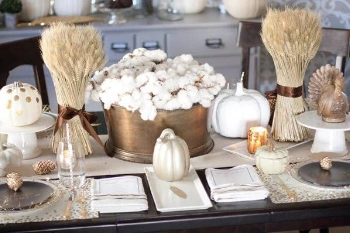 two bunches of dried wheat stalks, tied with brown ribbons, and placed in clear vases, on either side of a brass pot, filled with cotton branches, thanksgiving table ideas