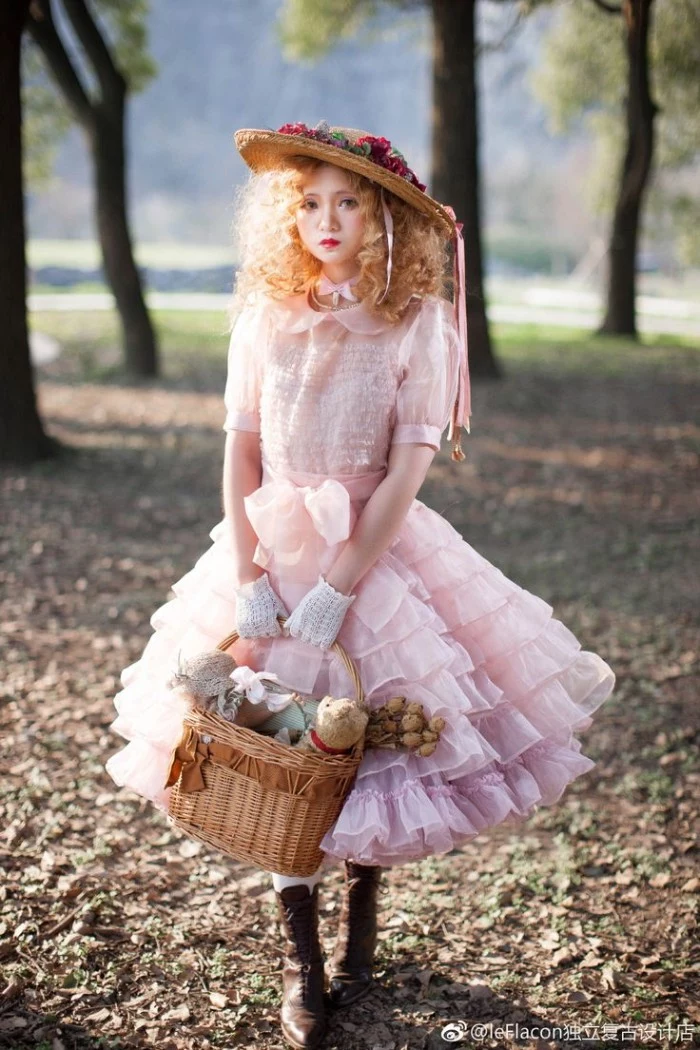 doll-like lolita style outfit, featuring a voluminous, frilly pale pink dress, a straw hat decorated with faux flowers, and a basket bag, worn by a woman with a curly ginger wig, and exaggerated makeup
