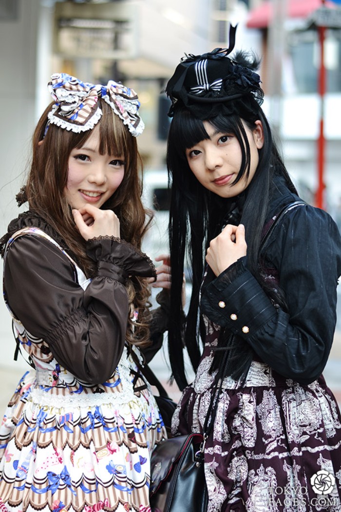 two girls dressed in gothic lolita dresses, brown and black shirts, worn under patterned pinafores, a large bow and a black hat 