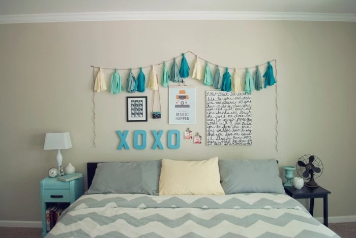 garland with cream, pale blue and turquoise tassels, hanging on a cream wall, decorated with three posters, and other ornaments, bed with three pillows nearby