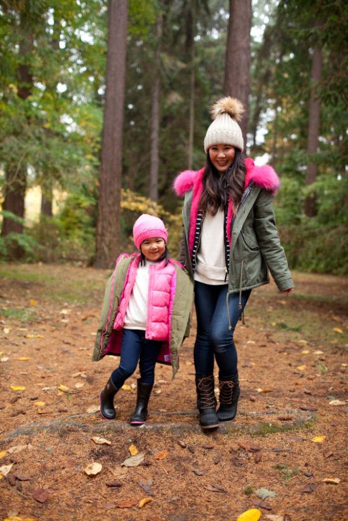 matching outfits for moms and daughters, skinny dark blue jeans, white jumpers and pale green parkas, toddler thanksgiving outfit ideas, on a little girl, standing next to her mom, in the forest
