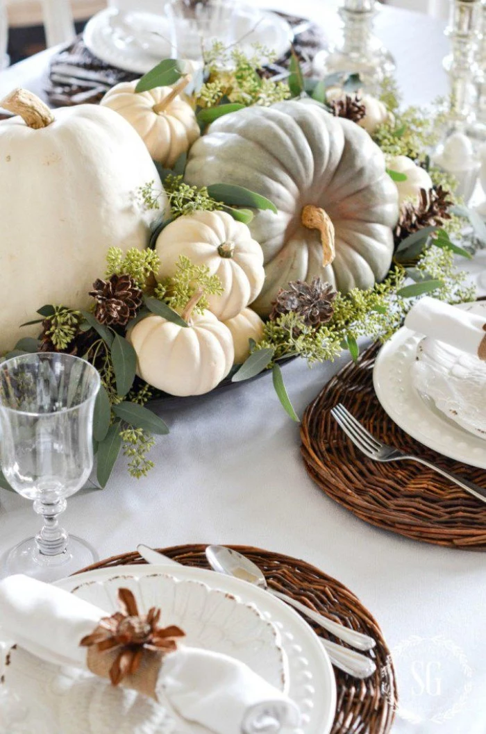 light grey and cream pumpkins, surrounded by purple, and pale green plants, on a table with round, brown wicker table mats, white dishes and napkins, glasses and cutlery, cheap centerpiece ideas