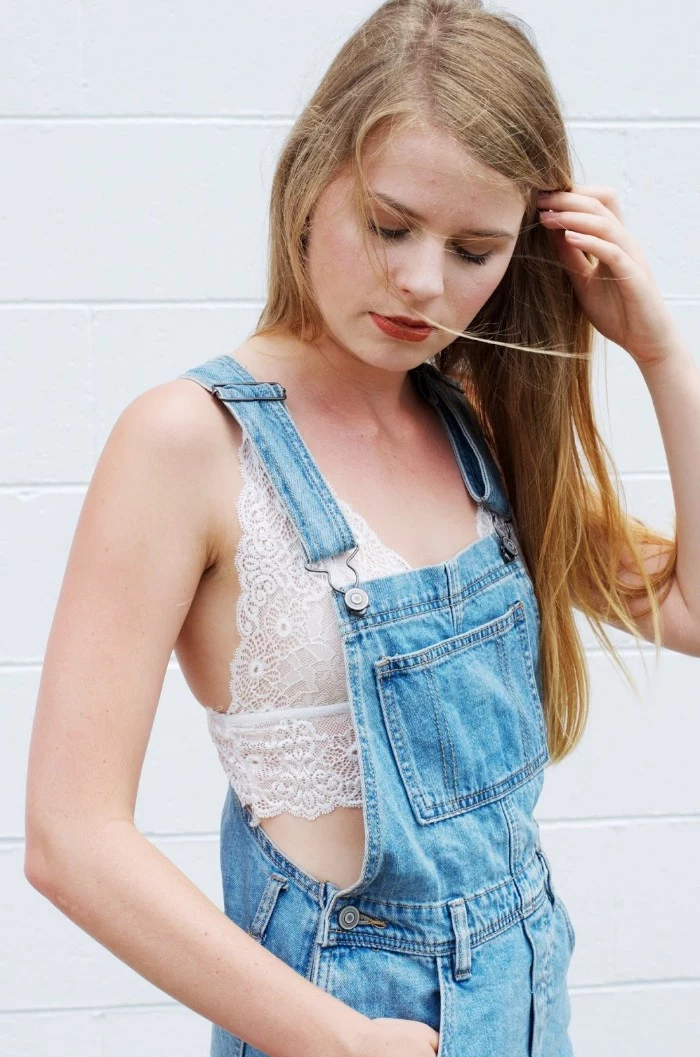 overalls made from light blue denim, worn over a white lace bralette, by a young woman, with dark blonde hair, bralette outfit in a casual style