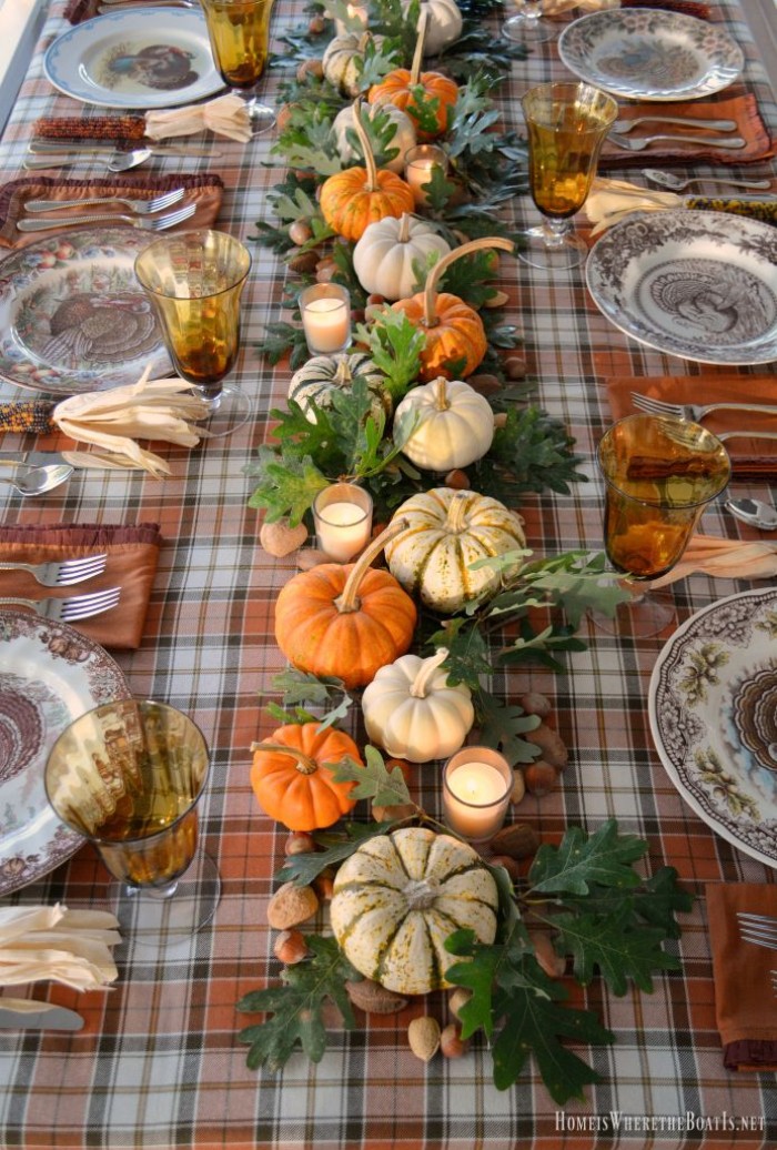 plaid tablecloth in orange, brown and white, on a rectangular table, decorated with orange, white and cream pumpkins, and dark green leaves, thanksgiving tablescape, ornamental plates and borwn napkins