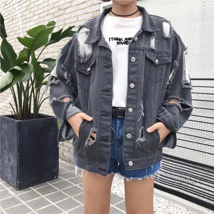 ripped oversized grey denim jacket, with many holes, 90s grunge fashion, worn by a slim woman, in a white t-shirt with black print, and blue denim cutoff shorts