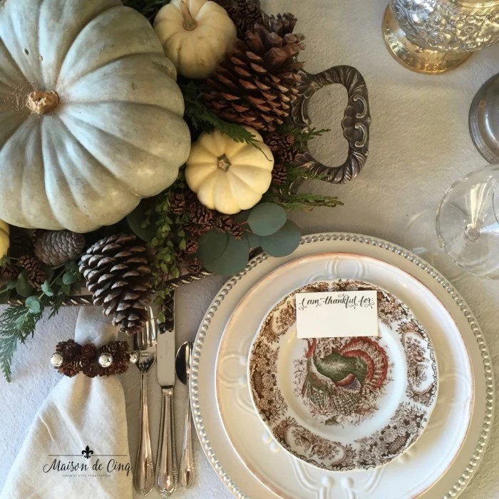 pinecones in different sizes, a pale duck's egg blue pumpkin, surrounded by small white pumpkins, and green leaves, on an ornamental silver dish, thanksgiving centerpiece, placed near three stacked dishes, silver cutlery and a whte napkin 