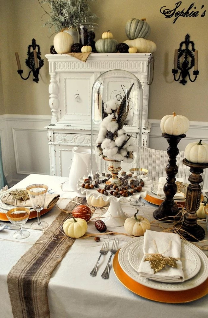 branch of cotton, and brown spotted feathers, in a white and gold vase, placed on top of a white dish containing acorns, thanksgiving centerpiece, pumpkins and a pinecone nearby