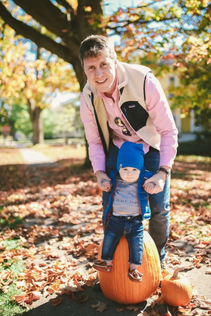 man dressed in a light pink shirt, dark blue jeans, and a pale cream gilet, holding the hands of a small baby, wearing dark blue jeans, a grey t-shirt, blue quilted jacket, and a blue beanie hat, baby thanksgiving outfits for little boys