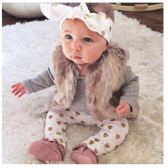 stylish baby girl thanksgiving outfit, white trousers with gold hearts, pale grey jumper with long sleeves, faux fur gilet, and a white and gold headband