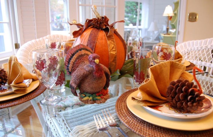 bird figurine in brown and orange, on a glass table, with plates and a large pumpkin, tied with ribbons, turkey decorations made from pinecones, orange pipe cleaners, and yellow napkins