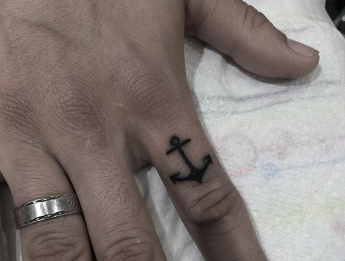 close up of a hand, with a silver ring on the ring finger, and a small black anchor tattoo, on the forefinger