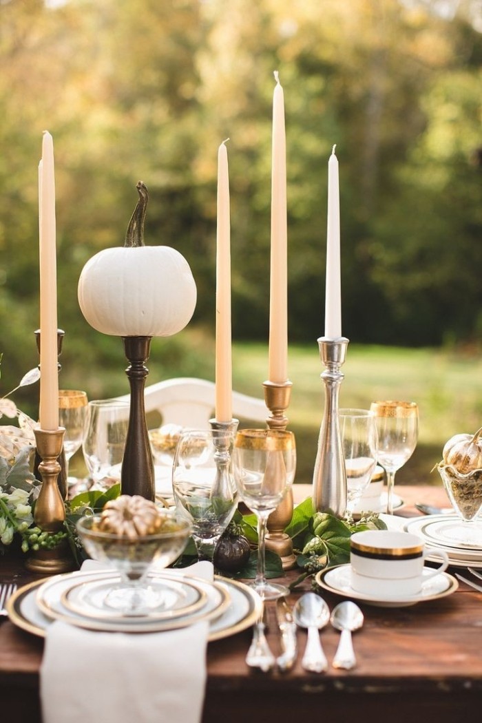boho style thanksgiving dinnerware, gold-rimmed dishes and glasses, tall cream and white candles, on a dark brown wooden table, decorated with pumpkin ornaments, and placed in a garden 