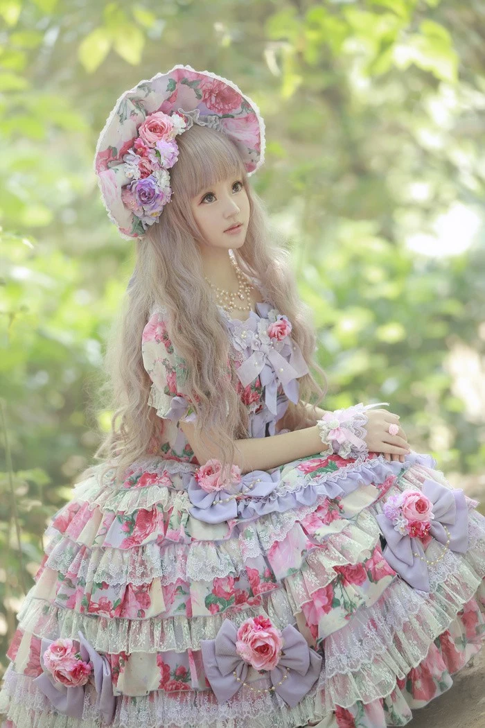 tiered dress with lace incerts and flounces, featuring a floral pattern, in pink and green, lolita fashion on a girl, wearing a florak bonnet, with faux flowers, and a long ash blond wig