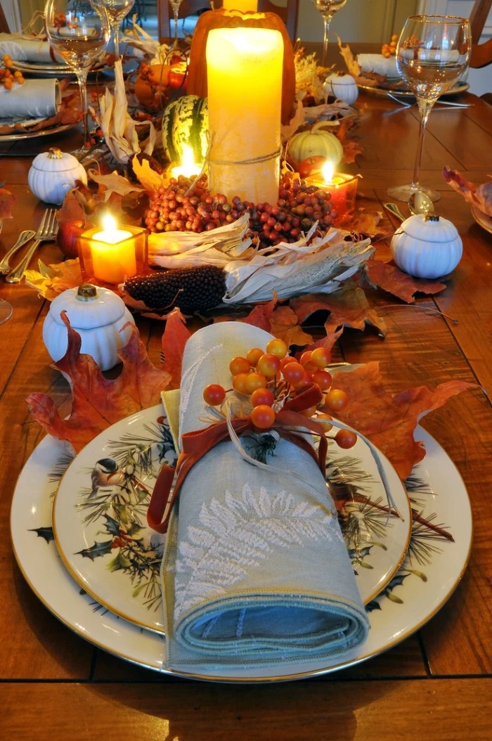 rolled up pale blue napkin, with a white botanical embroidery, tied with an orange ribbon, and decorated with small, yellow and orange berries, on top of two stacked plates, fall leaves and lit candles