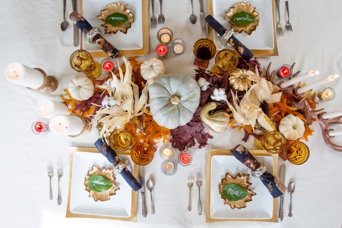 square yellow and white plates, with place holders, on a white tablecloth, decorated with a pale, duck's egg blue pumpkin, thanksgiving centerpiece, surrounded by fall leaves, dried corn leaves, candles and smaller white pumpkins