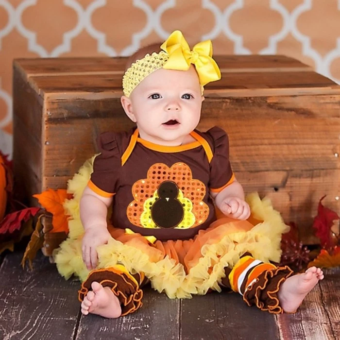 headband with a big yellow bow, worn by a small baby, dressed in a brown, orange and yellow dress, with a sequined turkey motif, and a frilly tutu, baby girl thanksgiving outfit ideas