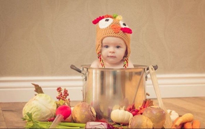 metal cooking pot, containing a small baby, wearing a turkey knitted cap, baby thanksgiving outfits, cabbage and carrots, and other vegetables near the pot