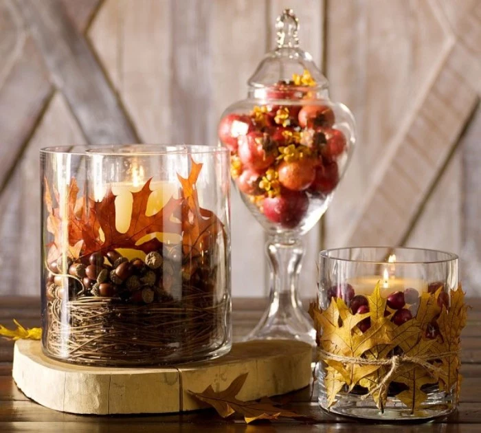 acorns and fruit, fall leaves and nuts, and lit candles, in and around three glass containers, placed on a dark brown table