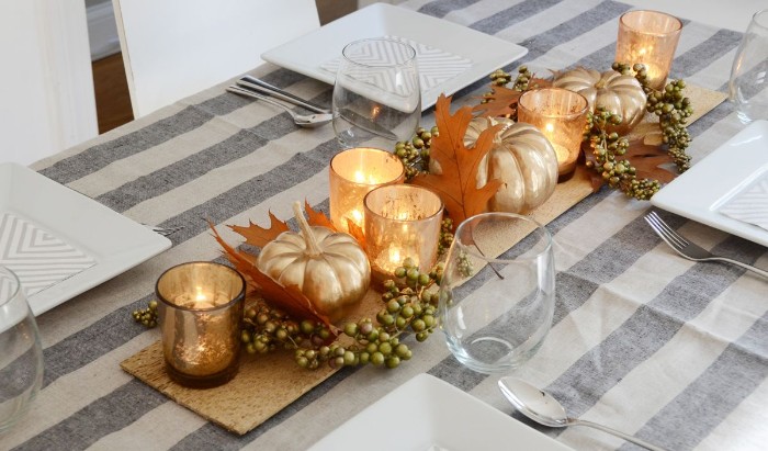 spray-painted faux pumpkins, in metallic gold, surrounded by tea lights, orange fall leaves and small green berries, thanksgiving centerpiece, on a grey and off-white tablecloth, with large square white plates